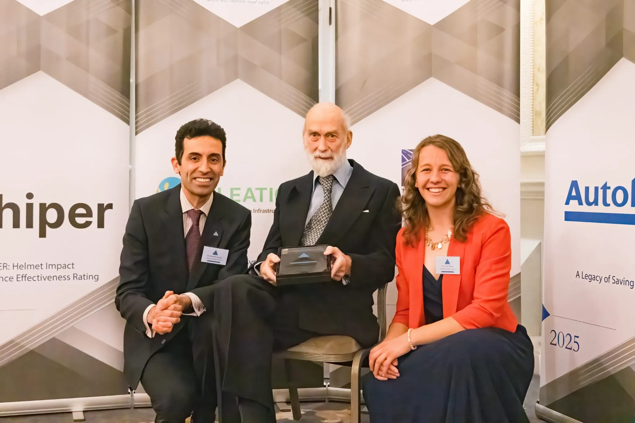 Professor Mazdak Ghajari and Dr Claire Baker with HRH Prince Michael of Kent, holding the 2025 Prince Michael International Road Safety Award, in front of a HIPER banner.